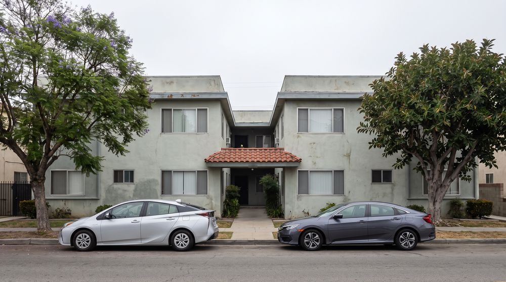 A two-story stucco courtyard apartment building in Los Angeles seen from across the street on an overcast day, with a flat parapet roof, a clay tile canopy over the entry and two parked cars