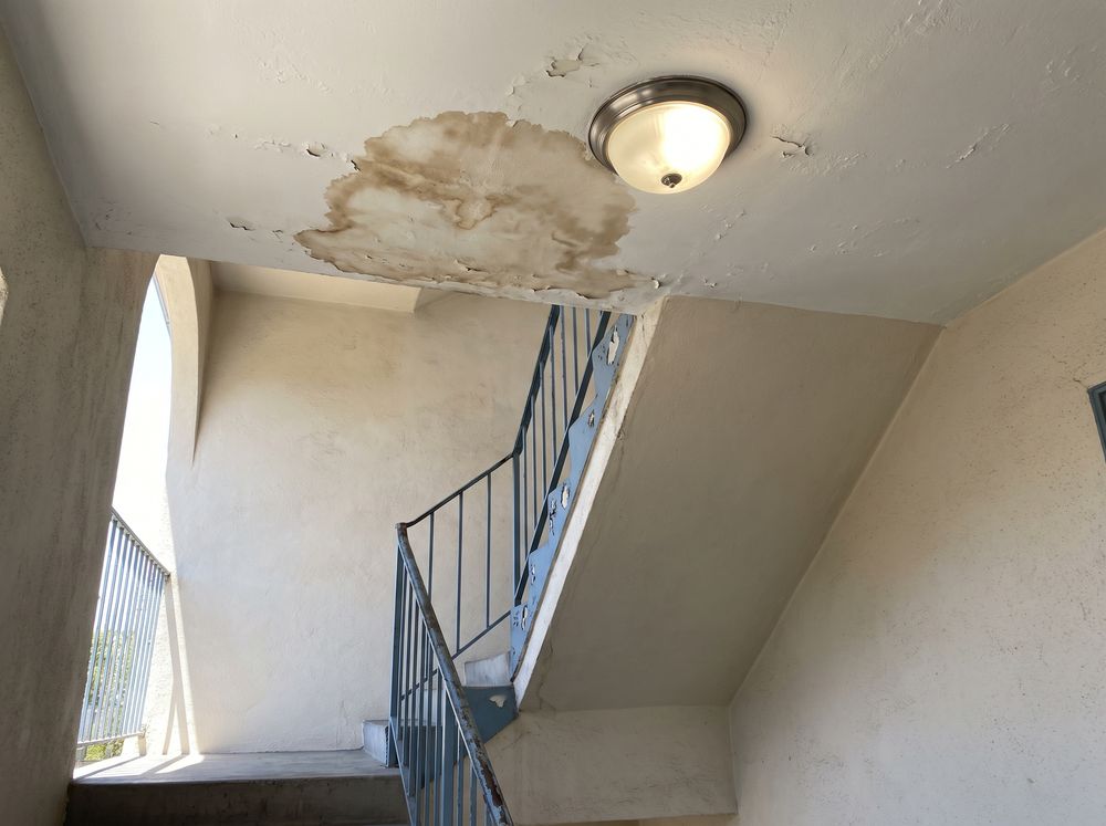 A brown water stain on the plaster ceiling of an open stairwell landing, beside a lit ceiling light fixture