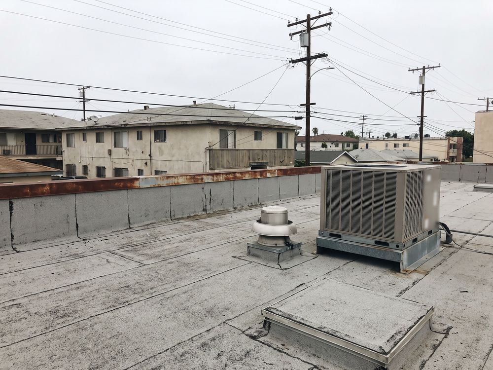 The low-slope cap sheet roof of a one-story row of shops in Los Angeles, with a rooftop air conditioning unit, an exhaust fan, a roof hatch and a parapet