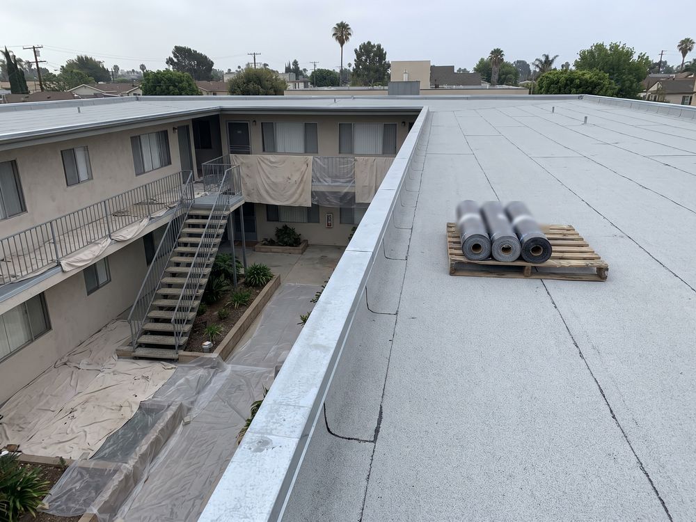 The flat roof of a two-story walk-up apartment building in Los Angeles in new light gray cap sheet, with rolls of cap sheet on a pallet and the courtyard below under drop cloths and plastic sheeting